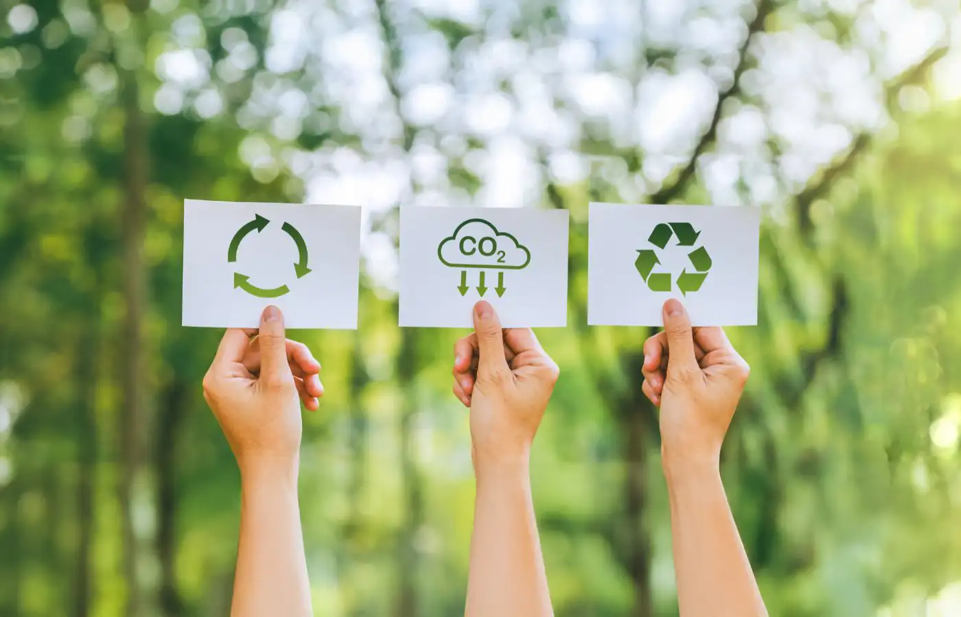 Trois mains tenant des cartes avec des icônes représentant le recyclage, la réduction des émissions de CO2 et la durabilité, sur un fond naturel.